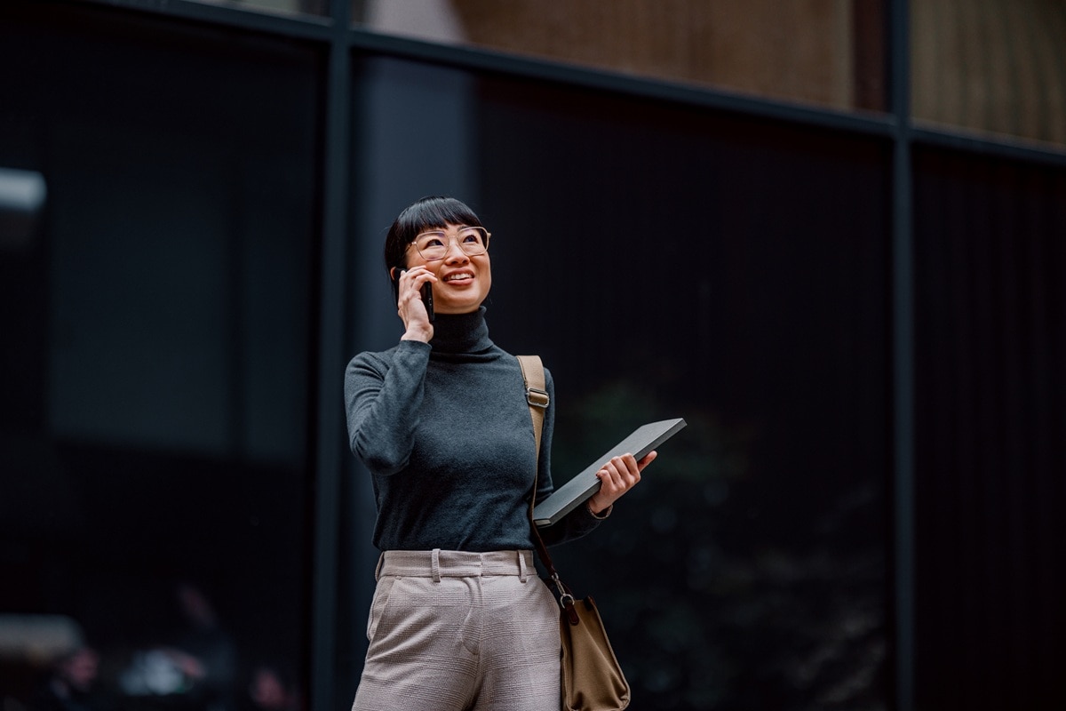 lady wearing glasses and black jumper walking with laptop on phone