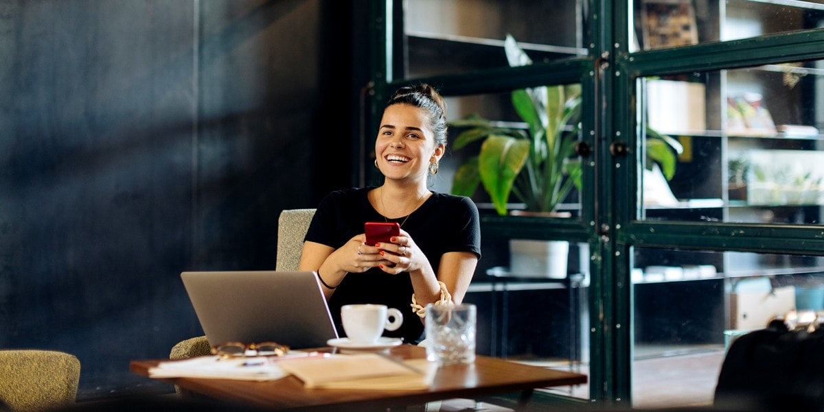 woman sitting at a desk holding cell phone, in the office, laptop and coffee mug in front of her